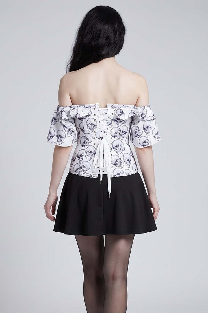 A model shown from the back wearing an Luxe Noir Corset off-the-shoulder white top with a black skull pattern and a black pleated skirt, set against a plain light gray studio background.