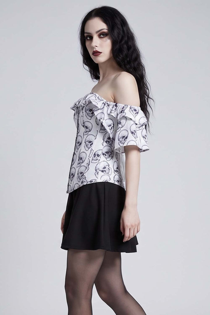 A woman modeling an Luxe Noir Corset off-the-shoulder blouse with a white base and black skull pattern, paired with a black skirt and sheer black tights, against a light gray studio background.