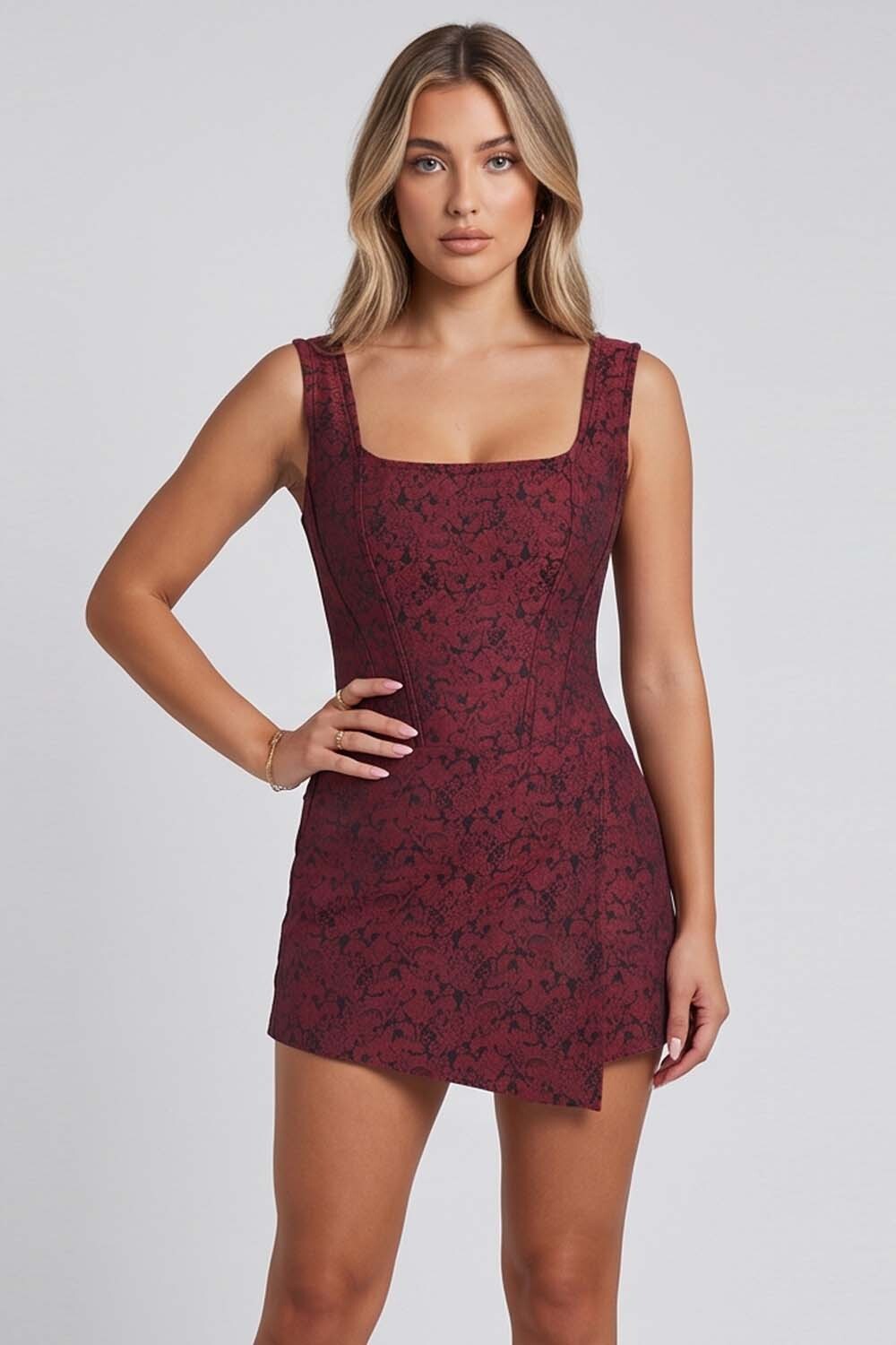 A woman modeling a sleeveless, scoop-neck mini Corset Skort dress with a dark red and black floral lace pattern against a light gray studio background.