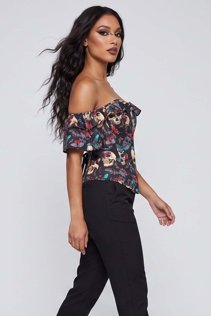 A woman modeling an Luxe Noir off-the-shoulder ruffled top with a dark multicolor skull and floral print, paired with black pants, against a light gray studio background.